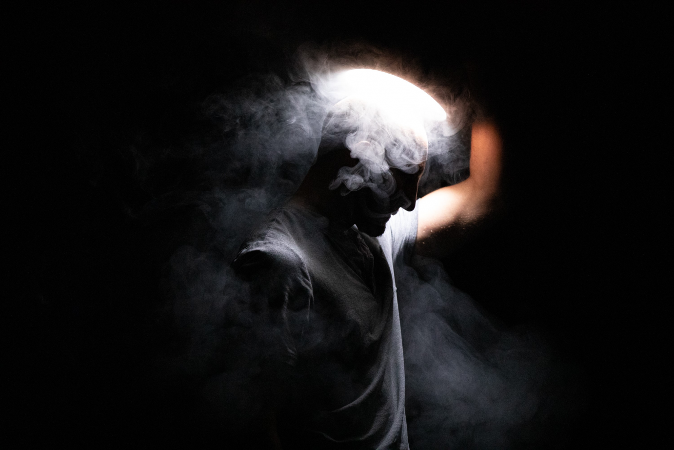 A man stands in profile, holding a light over his head as fog emits.